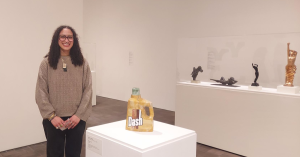 A person stands in a museum next to a sculpture resembling a yellow Dash detergent bottle. Several other sculptures are displayed on white