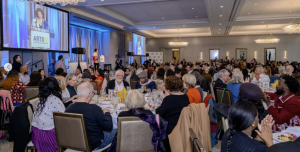 A large group of people sit at round tables in a banquet hall, listening to a speaker at a podium. The event is formal, with a projected scr