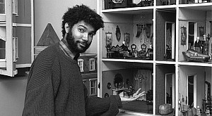 A young Fred Wilson, with curly hair and beard, wearing a sweater, gestures toward an elaborate multi-room dollhouse filled with furniture