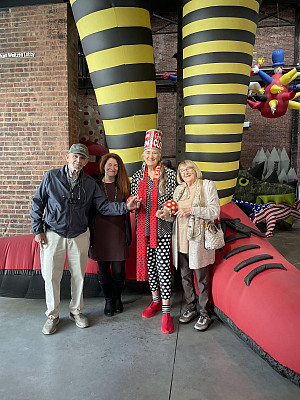 Four adults stand smiling in front of a whimsical, oversized inflatable sculpture with black and yellow striped legs. The group holds hands,