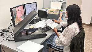 Student with dark long hair, white blouse, and jeans sits at an office desk with a book, desktop computer, and scanner.