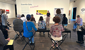 A group of museum visitors sits facing a wall with artwork, listening to a guided tour