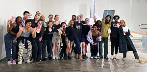 A group of dance students standing side-by-side in a museum gallery with pieces of an artwork visible in the background