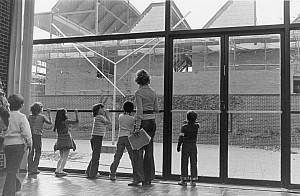 Black and white archival image of children and a teacher looking through large windows at a brick building under construction