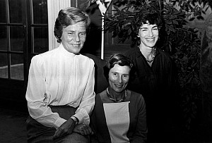 Archival black and white image of three women in seated positions facing the camera and smiling