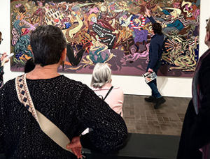 A male curator gestures to a detail in a large, colorful artwork while a group of adults gather around to listen