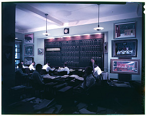 Archival color photograph of an office with gentlemen working at rows of desks facing a Wall Street-style trading board flanked by artworks.