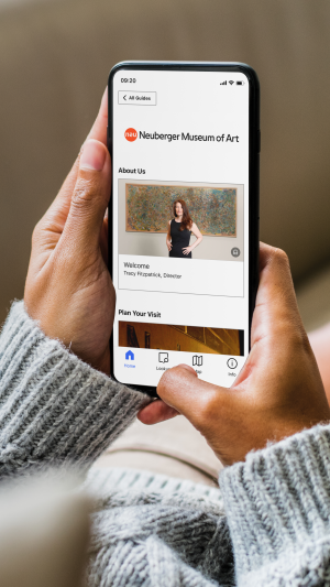 A person holds a smartphone displaying the Neuberger Museum of Art app, showing a welcome message from the director and navigation icons at
