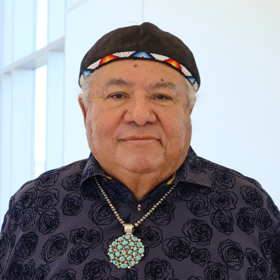 An older man stands indoors wearing a patterned dark shirt, a beaded cap, and a large turquoise necklace, with his hands behind his back and