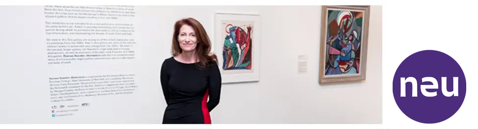 Museum Director Tracy Fitzpatrick, a white woman with brown hair wearing a black dress standing in a gallery with colorful artwork