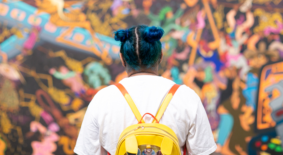Photo of the back and shoulders of a college aged female wearing a backpack and with blue hair looking at a large piece of art