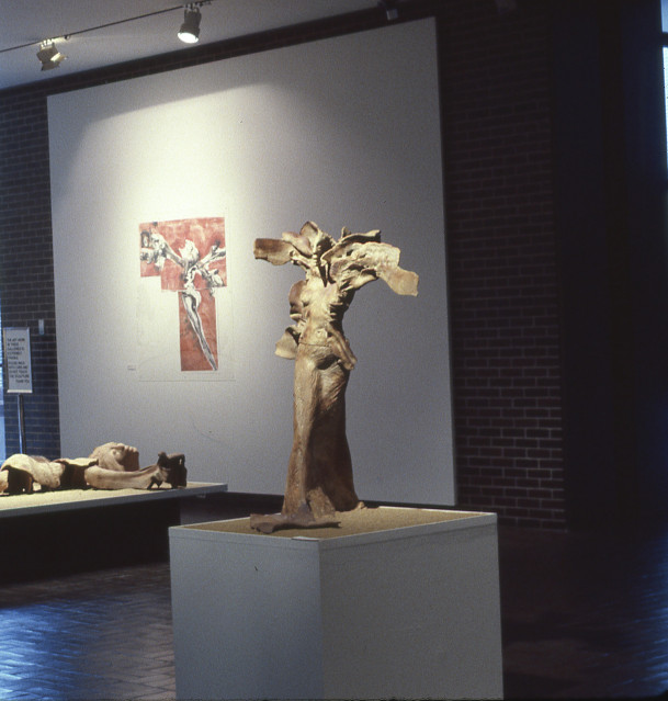 An installation photograph of a beige 3D representation of a female figure. Around her head are slabs of ceramic that could be floral or ...