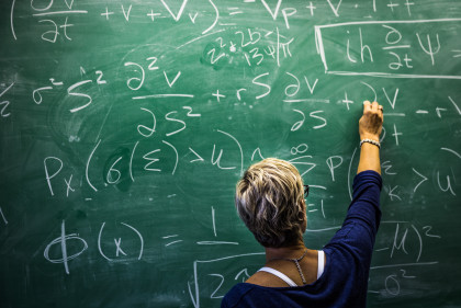 A teacher writing Mathematics equations on a chalk board of a classroom.