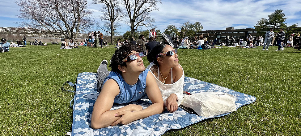 Two students laying in a blanket in the grass wearing eclipse glasses and looking up.