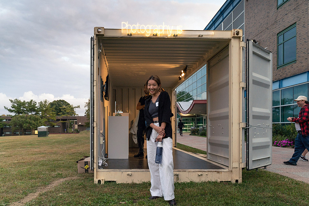 Student stands in front of an open shipping container with Photography Is in neon on top.