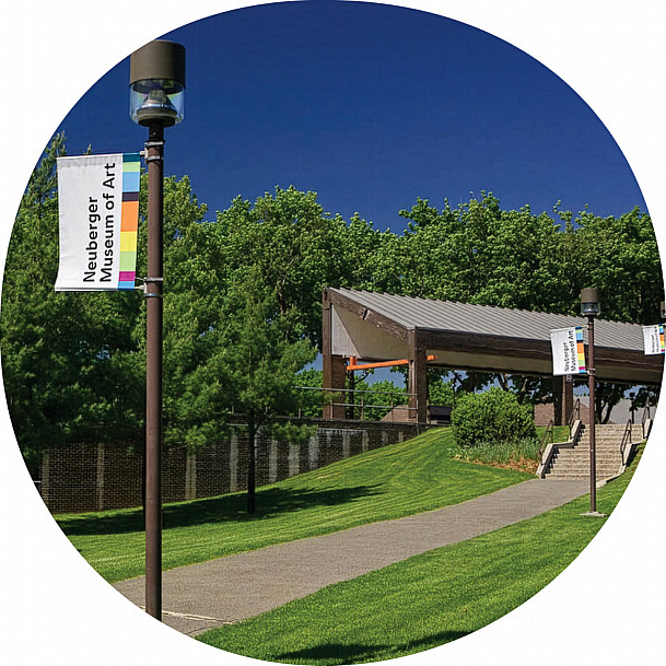 Pathway leading to the main campus with rainbow banners, trees, grassy slope, and modern pavilion under a clear blue sky.