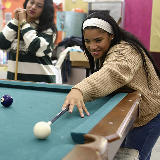 Student with tan sweater and white headband with a pool cue aims at the cue ball.