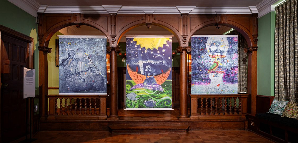 Three colorful prints on textiles hang in gallery with wood floors and walls.