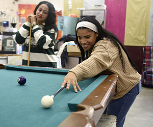 Student with tan sweater and white headband with a pool cue aims at the cue ball.
