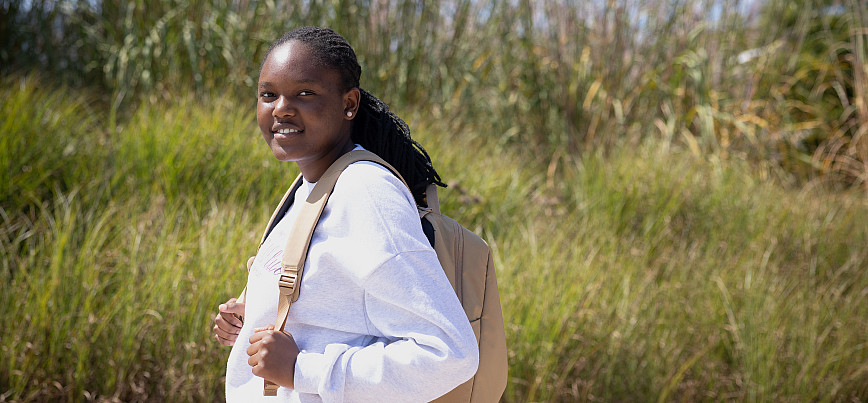 240912 PUR492 Student with backpack and white sweatshirt walking and smiling vegetation behind
