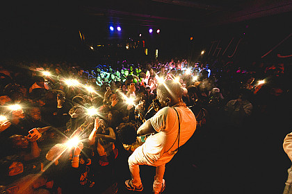 Performer fro behind in white on stage with blurry lights and crowd in front
