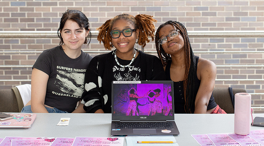 Three smiling students sit behind a table