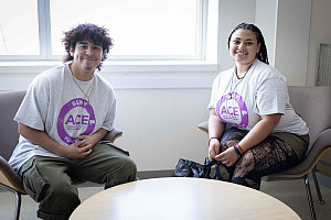 Two students wearing white t-shirts with purple ACE logo sit in chairs, smiling.