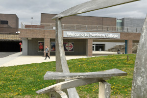 Person walks in distance with Welcome to Purchase College sign on brick building