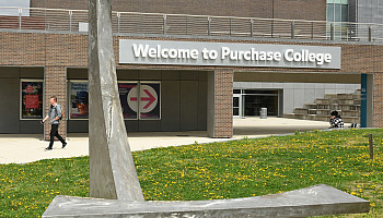 Person walks in distance with Welcome to Purchase College sign on brick building