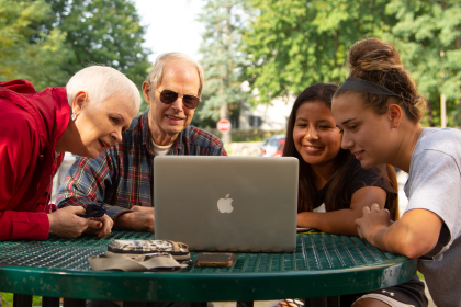 Senior citizens and college students outdoors sitting at table, looking at laptop