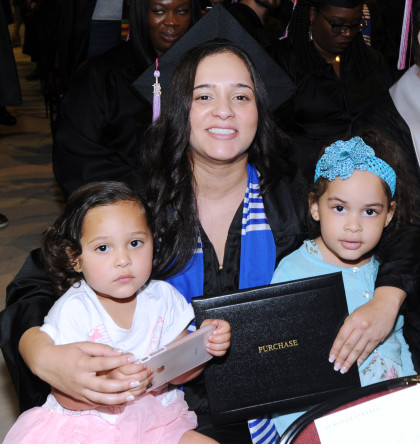 Graduate in cap and gown embraces two young children holding a diploma cover.
