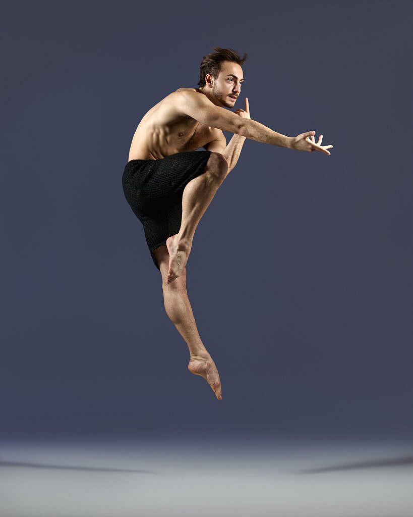 Dancer Deniz Erkan Sancak leaping with one knee bent and the other extended down.