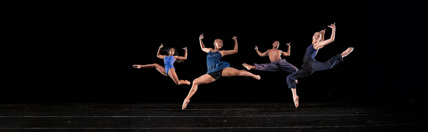 Four dancers leap in unison on a dark background.