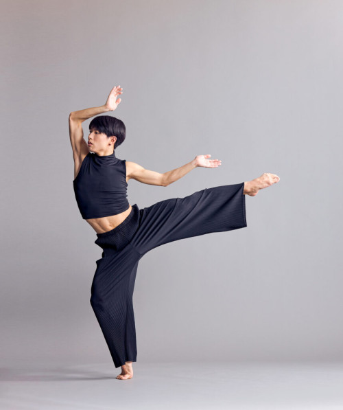 Asian dancer dressed in black with leg up and arms overhead at angles.