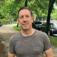 Graham Rayman in a gray shirt standing on a path of trees.