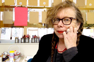 face of woman with glasses, red lipstick and two long braids in front of art supplies