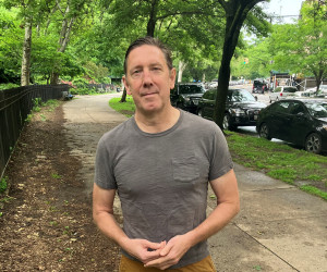 Graham Rayman in a gray shirt standing on a path of trees.