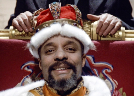 Robert Liensol as The Parliamentarian in close-up sitting on a throne with a crown on.