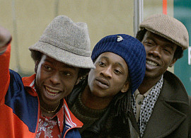 Three men lined up behind each other look towards the bottom left of the screen. The guy in the front points off in the same direction.