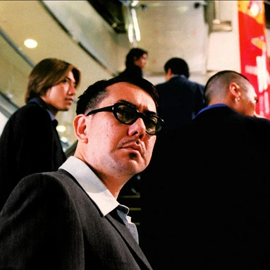 The five main characters of the film ascending up an escalator at a mall. One character in the foreground looks behind them.