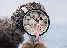 Mug of hot chocolate with marshmallows and candy cane being held by a hand with a mitten.