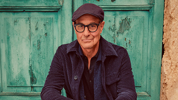 Stanley Tucci in a hat and glasses in front of a teal green door.