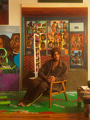Artist sits in a chair surrounded by brightly colored narrative paintings