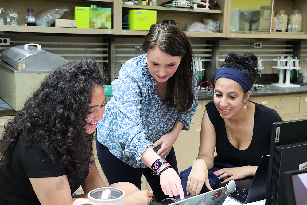 Professor stands over two students seated at laptops and points at a screen.