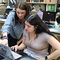 Professor stands next to student sitting at laptop in a science lab.