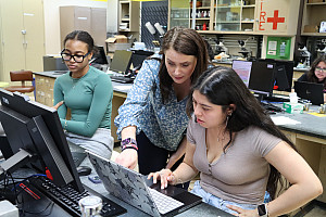 Professor stands next to student sitting at laptop in a science lab.