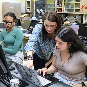 Professor stands next to student sitting at laptop in a science lab.
