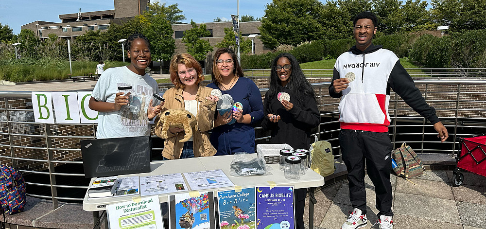 Five students smiling and standing behind a table on the campus plaza