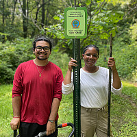 Two smiling interns with tools stand by a nature preserve sign on a wooded trail surrounded by green trees and grass.