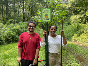 Two smiling interns with tools stand by a nature preserve sign on a wooded trail surrounded by green trees and grass.